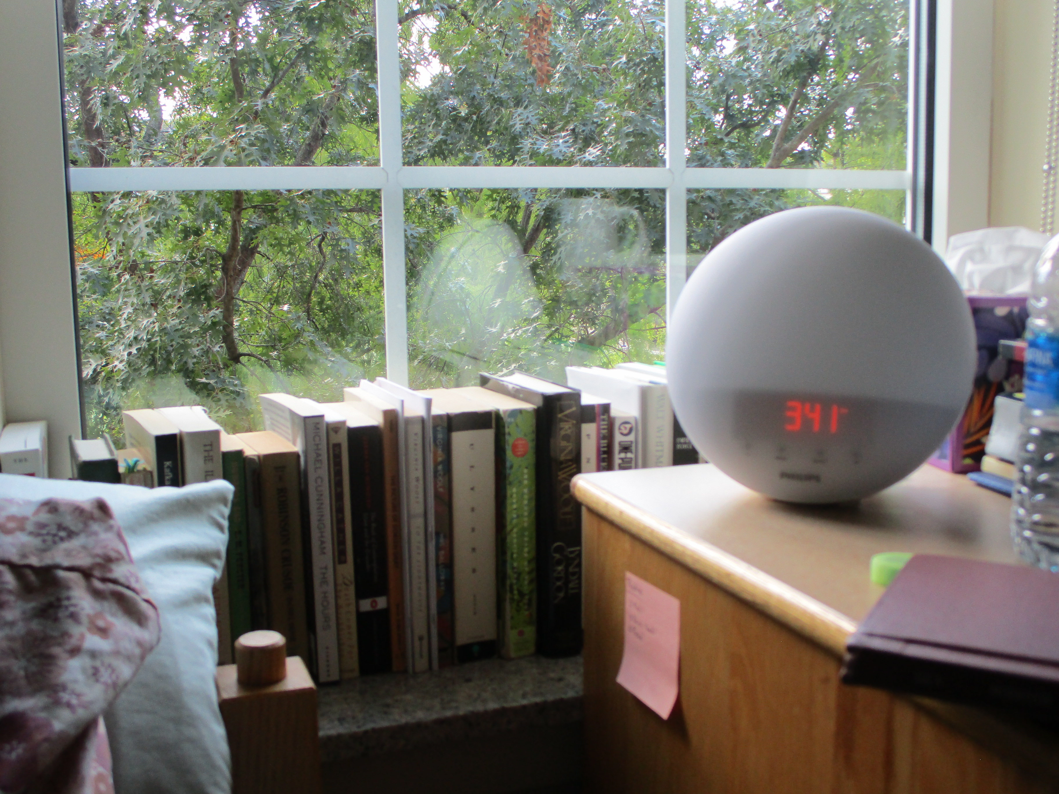 A picture of my bedside: pillows, a windowsill lined with books, and a sunrise lamp.