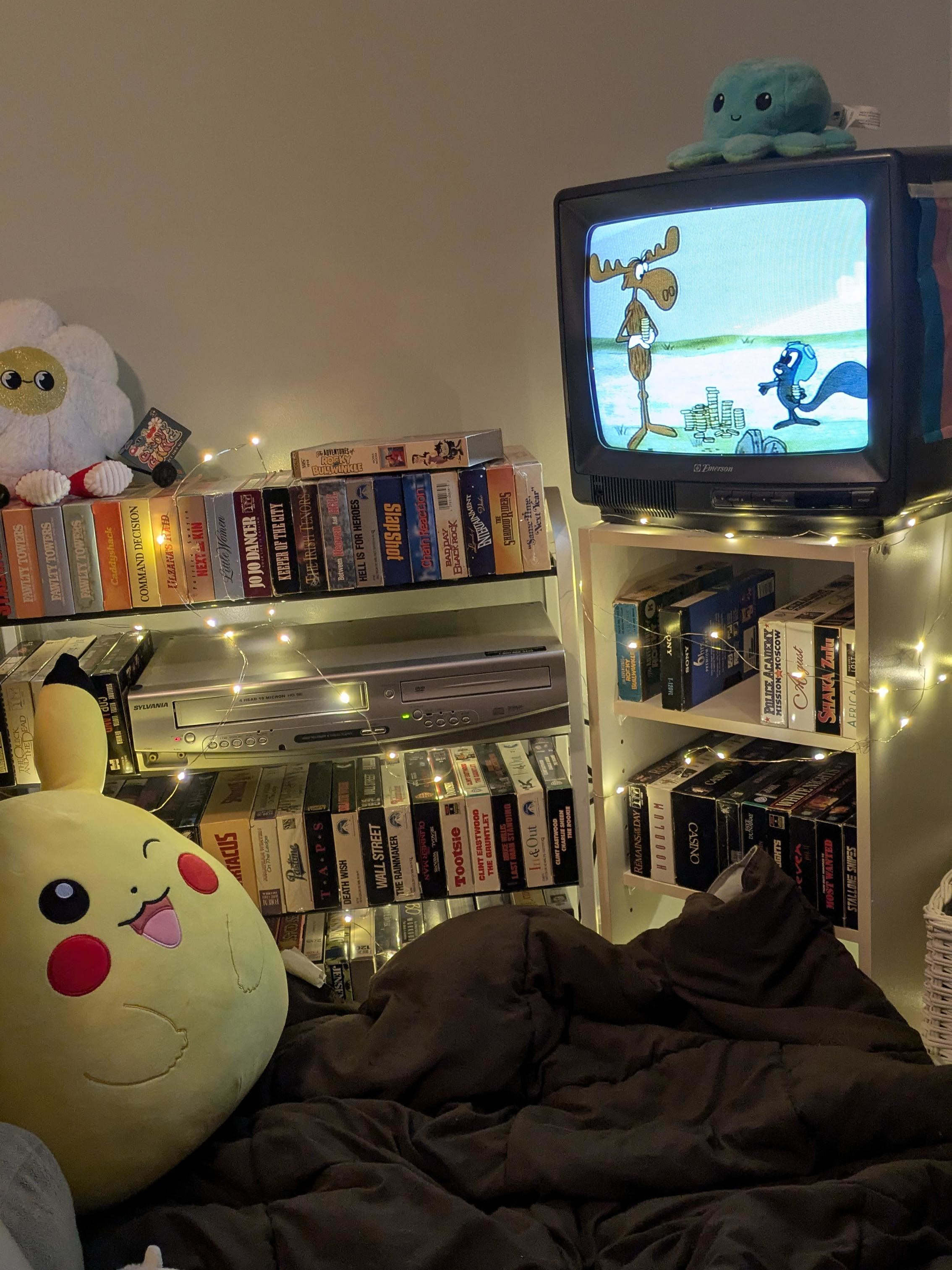 A warmly lit corner of two bookshelves of vhs tapes. On top of one sits a CRT television which is playing Rocky and Bullwinkle. There is a Pikachu plush on the ground.